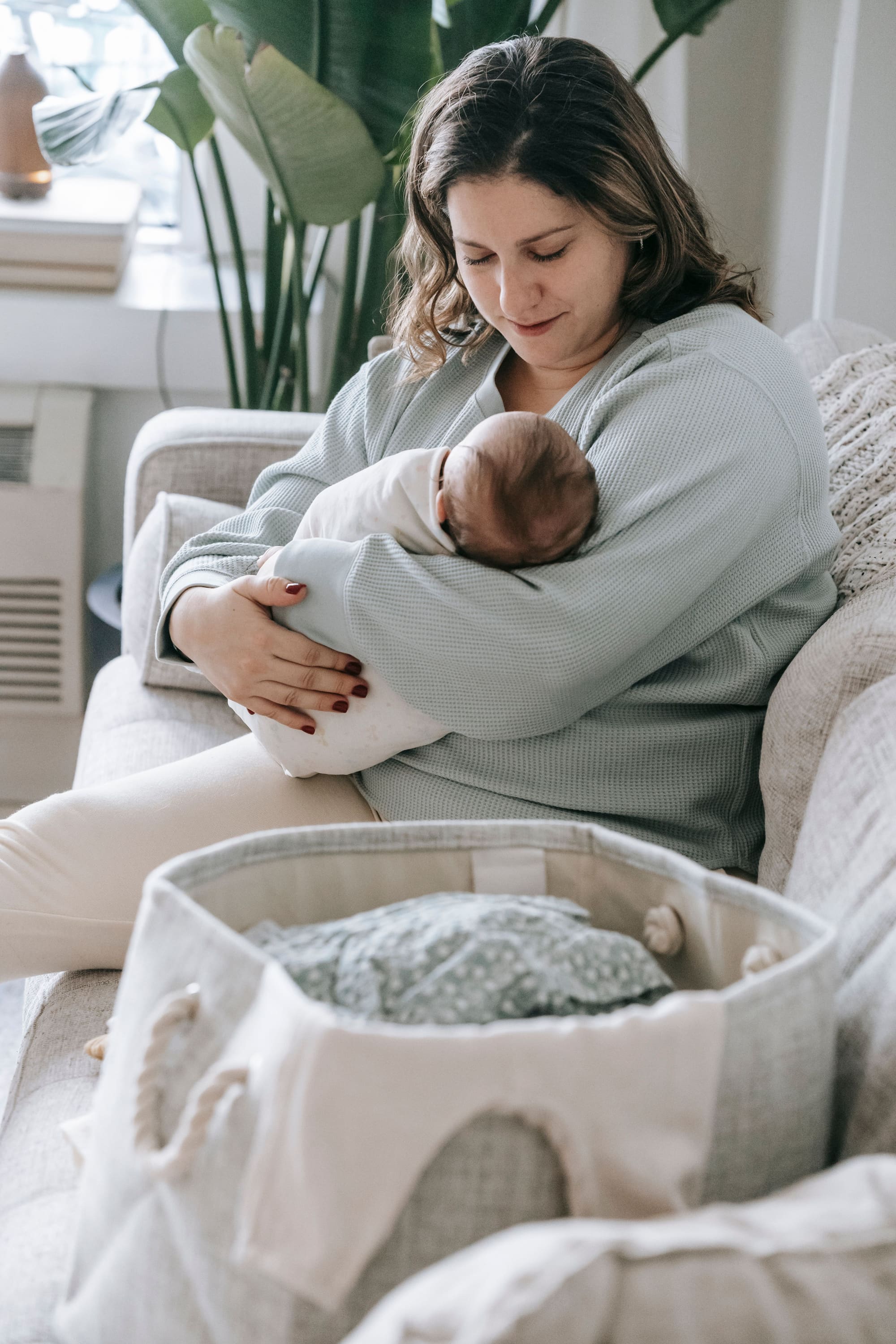 Caring mother with little infant baby in arms sitting on comfortable sofa with basket of clothes in living room at home